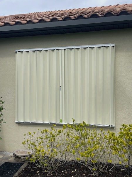 Beige Eurex Shutters PTX Accordion Shutters on medium sized window of a beige house in the closed position.