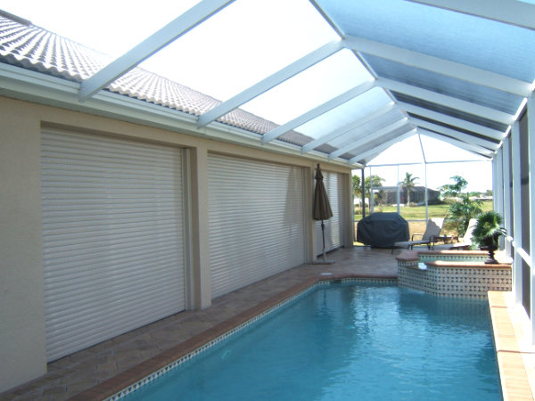 White roll-down shutters shown installed over the openings of a lanai near a blue pool