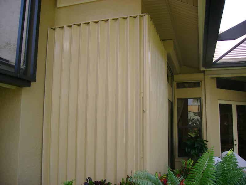 Beige accordion shutters shown both open and closed over the front windows of a beige home in Florida