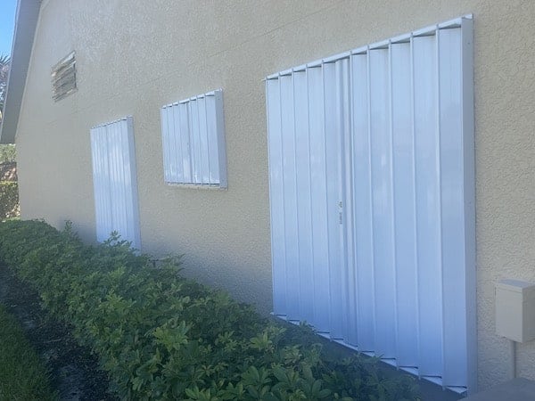 Three white colored accordion shutters shown closed over three different sized windows on a home in Punta Gorda Florida.