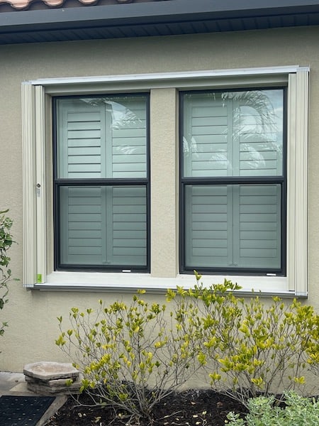 Beige accordion shutters with locks on a beige home. Shown in the fully open position. 
