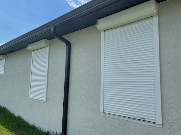 white rolling shutters in the closed position covering windows of a tan home