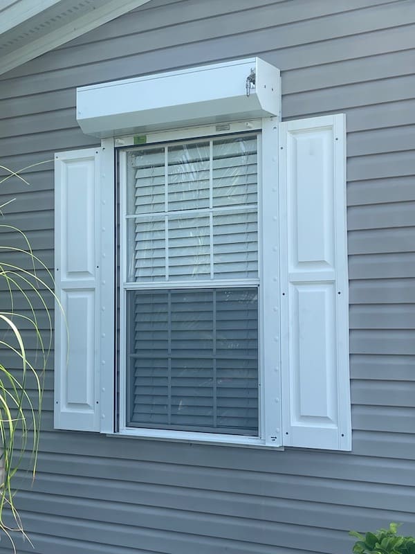White, open roll shutter installed over a single window on a beige home.